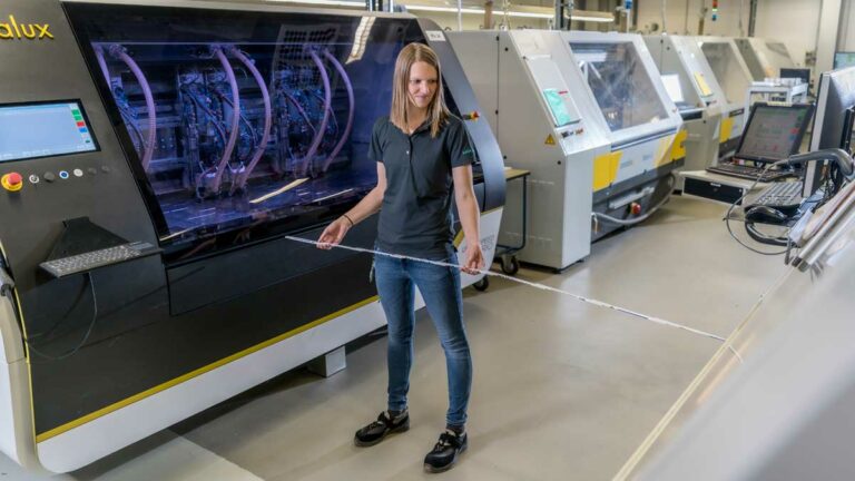 Anne Heinig stands between machines for PCB production and holds a folding rule in her hand with which she measures the distance.